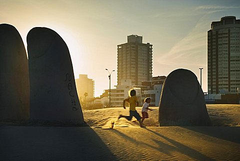 La Mano Sculpture of a hand on the beach at Praya Brava with children playing in the evening light, Punte del Este, Uruguay